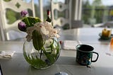 A white and purple flower arrangement in a round clear vase, in the morning light on a table next to a coffee mug. A spent wine bottle in the background.