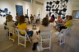A group of people sits in an art gallery in a circle in white wood folding chairs. In the center is a microphone sitting on a small table. The backdrop is an art gallery with a variety of blue works on the walls. The photo is taken from behind so we see the backs of participants in the foreground and those facing the camera are further away.