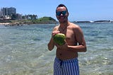 The author is shirtless and wearing sunglasses, a white swim short with blue stripes and a neckless. He is holding a coconut while looking at the camera and smiling. He is at the beach and the water is crystal clear blue water. All the way in the back, you see a green hill and to the left, a hotel. The sky is clear as there are no clouds.