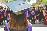 Photo of a person wearing a graduation cap taken from behind, facing a blurry crowd of people in graduating gowns.