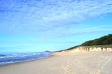 A long view of an empty sandy beach with footprints, the ocean, and a cloudy but magical brilliant blue sky.