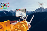 A person in a ski coat operating a drone over snow-covered mountains with an Olympic logo in the top-left corner.