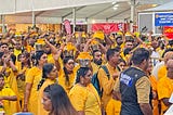 Large crowd of people wearing saffron colored clothing, some are carrying silver pots topped with yellow cloth, on their heads.