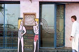 A colorful cartoon female & male in their swimming suit illustration on the menu board outside a seaside restaurant. The chef in his white uniform just arrived on the right-hand side of the photo, looking at the cartoon figures.
