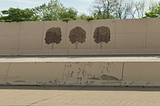 Concrete sound barrier on the side of a highway with three trees painted on it. You can also see a few trees growing behind the barrier that are somewhat taller than it is.