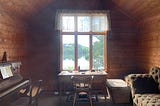 A small room with a piano and a desk with a stunning lake and nature view.