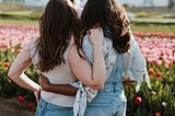 Two female friends with arms wrapped around each other looking out at a field of flowers