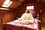 Woman sitting up in bed wrapped in a hand knit bulky blanket.