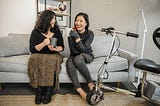 Two disabled women chat on a couch, both holding coffee mugs. An electric lightweight mobility scooter rests nearby.