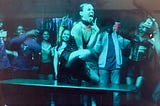 Quinni dancing on top of a table surrounded by drunken partygoers cheering her on. She is knelt down and sticking her tongue out with her fingers in a “V” formation just below. Her outfit is all black with a silk slip dress, a fishnet top underneath, fishnet tights, and black leather combat boots. Her red hair is slicked back and she looks exuberant.