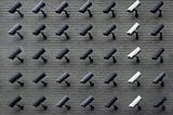 Many CCTV cameras that are differing shades of gray, mounted on a wall