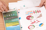 photo of a desk with someone working with paper showing graph, pie charts and a phone