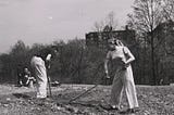 Black-and-white photo of two people tilling bare soil, preparing a garden plot. An apartment building is in the background.