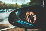 A side mirror of a car, reflecting the image of a woman aiming a digital camera towards the scenery on the side of the road.