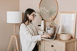 A young woman types at her computer, standing on her wooden drawer in her room beige, minmalistic, floral room.