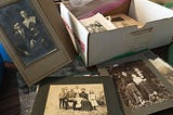 A worn cardboard box filled with old photographs sits on a wooden table in soft morning light, with several faded prints scattered nearby, suggesting reflection, memory, and the moments people hold closest over time.