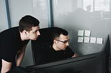 Two people in black shirts are working at a desk, one standing and peering over the shoulder of the other, who is seated on a chair. In the background, some sticky notes are posted on a glass wall.