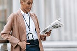 Smiling Black businesswoman reads newspaper outside modern building.