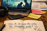 Laptop displays “Building Safer AI for Youth Mental Health Hackathon,” notebook reads “How do we make this safer for struggling youth?”