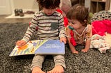 Two kids on a rug. The older child is reading to the younger child.