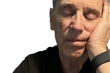 Older man with gray hair wearing a black shirt and smart wristband, resting his head on his hand with eyes closed, appearing to be asleep or deep in thought.