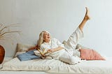 Woman lying in bed on her right side with a book in right hand. She’s looking away smiling joyfully with her left leg kicked up, pointing to the ceiling