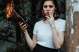 A woman holding a bottle with fire coming out of the top in one hand and a cigarette in the other, standing against a backdrop of plants and a weathered wall.
