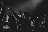 Black and white photo of a young person pointing at a band on a stage with a light overhead coming in from the heavens above.