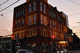 A Romanesque Revival style building at dusk at a major intersection with cars passing through.