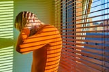 Distressed man with hands on head wearing an orange long-sleeved shirt standing by a window with venetian blinds