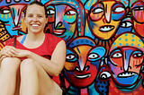 A woman smiles in front of a painted mural of many colorful faces