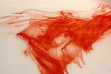 An image of red dye dropped in water that looks like a thread of blood, with ribbons of red color moving in waves around the thread.