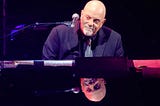 Billy Joel sitting a piano, smiling slightly and looking to the side. His face is reflected in the piano’s surface.