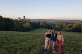 Eden and two female friends stand at the top of a grass hill in Durham. Behind them in the distance you can see the skyline of the city, along with the Cathedral. The sun is just beginning to set and the sky has a haze of pink and orange alongside blue. There is not a cloud in the sky.