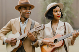 A black couple playing guitars dressed in 1920s clothing.