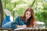 A girl lying on table in park writing.