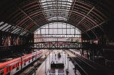 A train station that has a red train stopped on the left side, and some people waiting to board.