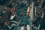 A vertigo-inducing first-person perspective looking down from an extremely high skyscraper or tower structure. The image shows someone’s feet in white sneakers dangling over the edge of a metal platform or scaffolding, with the sprawling cityscape visible hundreds of feet below. City streets form a grid pattern with illuminated buildings and traffic lights creating a tapestry of urban life at night. The composition creates a dramatic sense of height and danger.