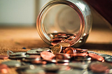 A glass jar tipped over spilling coins onto a flat surface. The view is looking directly inside the tipped over jar