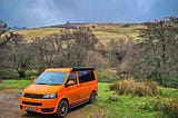 orange T5.1 Volkswagen campervan in the foreground with trees and a hill in the background