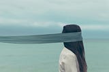 person facing the right with a wide teal ribbon covering their face and trailing far behind them out of the image, person’s head is covered in long, straight dark hair falling onto a white shirt, opaque sea or lake and cloudy sky in the background