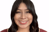 Photo of Yolis, a Hispanic woman, from the chest up. She is looking at the camera with an open-lipped smile. She is wearing a burgundy top. Her short brown curls are brushing both shoulders.