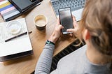 Person holding a mobile phone at a desk with a journal and coffee.