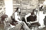 Three teenagers (two male and one female) sit on chairs in a bricked in room