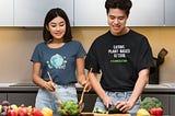 Millennial Asian couple in kitchen preparing a plant-based meal with green lettuce salad, red and yellow peppers plus broccoli, cucumbers, and tomato.