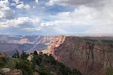 A view of the Grand Canyon with clouds in the sky above, trees in the foreground, and the river in the distance