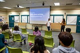 Ana Clara Otoni and Michael Lozano stand in front of a crowd of a dozen of teens in a library room presenting a slide show where it reads: “Media literacy workshop.”