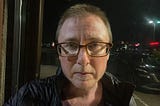 The author is a white man with short hair wearing glasses. He has a serious expression. It’s dark outside. A streetlight has lit up his face. He’s wearing a black jacket. He has stubble of a beard and mustache.