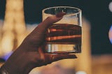 A woman’s hand with red nails holds a whiskey glass. The background is blurred, showing the lights of a city skyline, Las Vegas, at night.