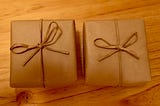 Two small gifts wrapped in brown paper and tied with brown paper strings into bows, sitting on a wooden surface.